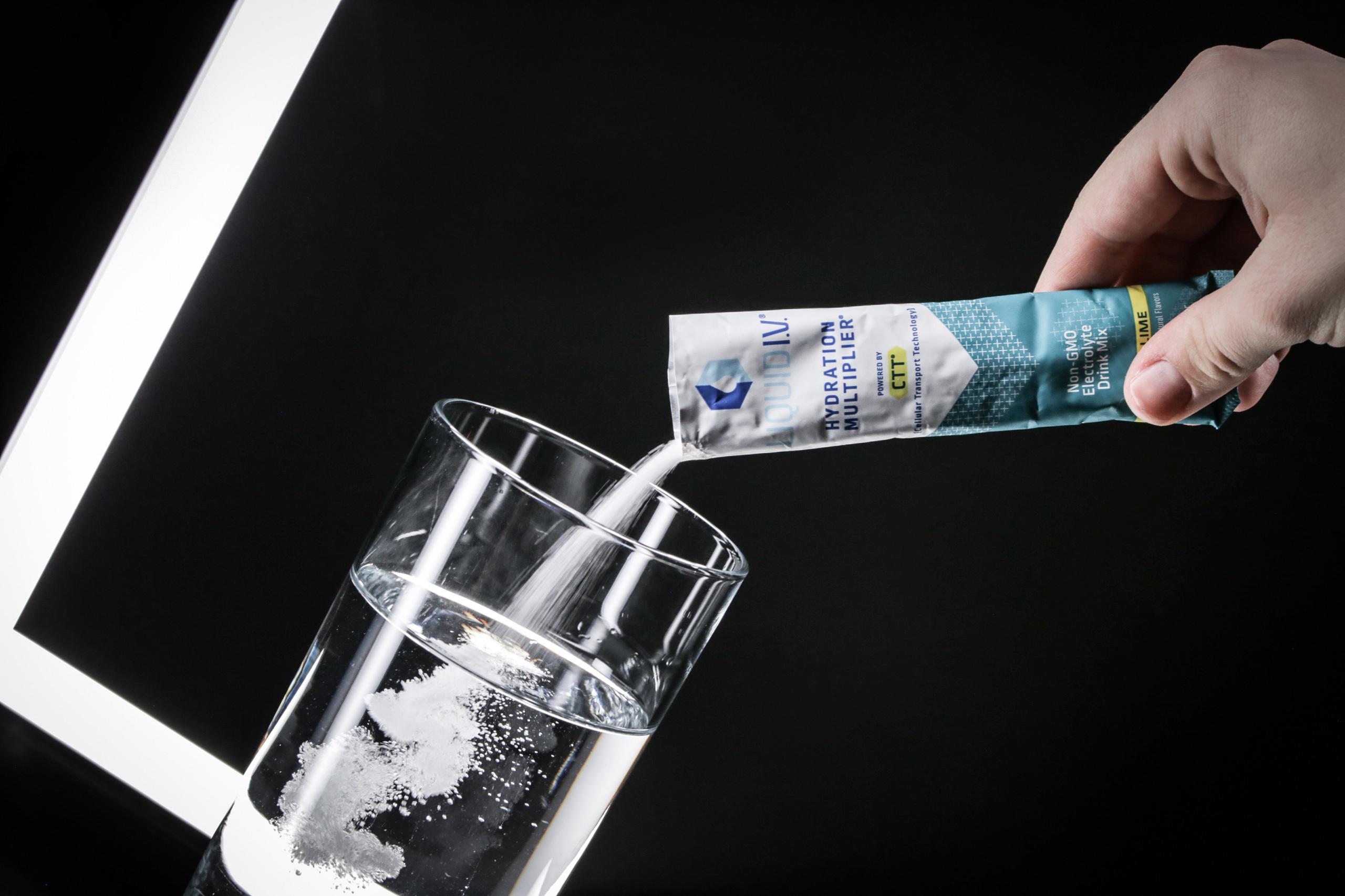 A hand squeezing a sachet labelled 'Liquid I.V. Hydration Multiplier' into a glass of water, with the powder visibly dissolving.