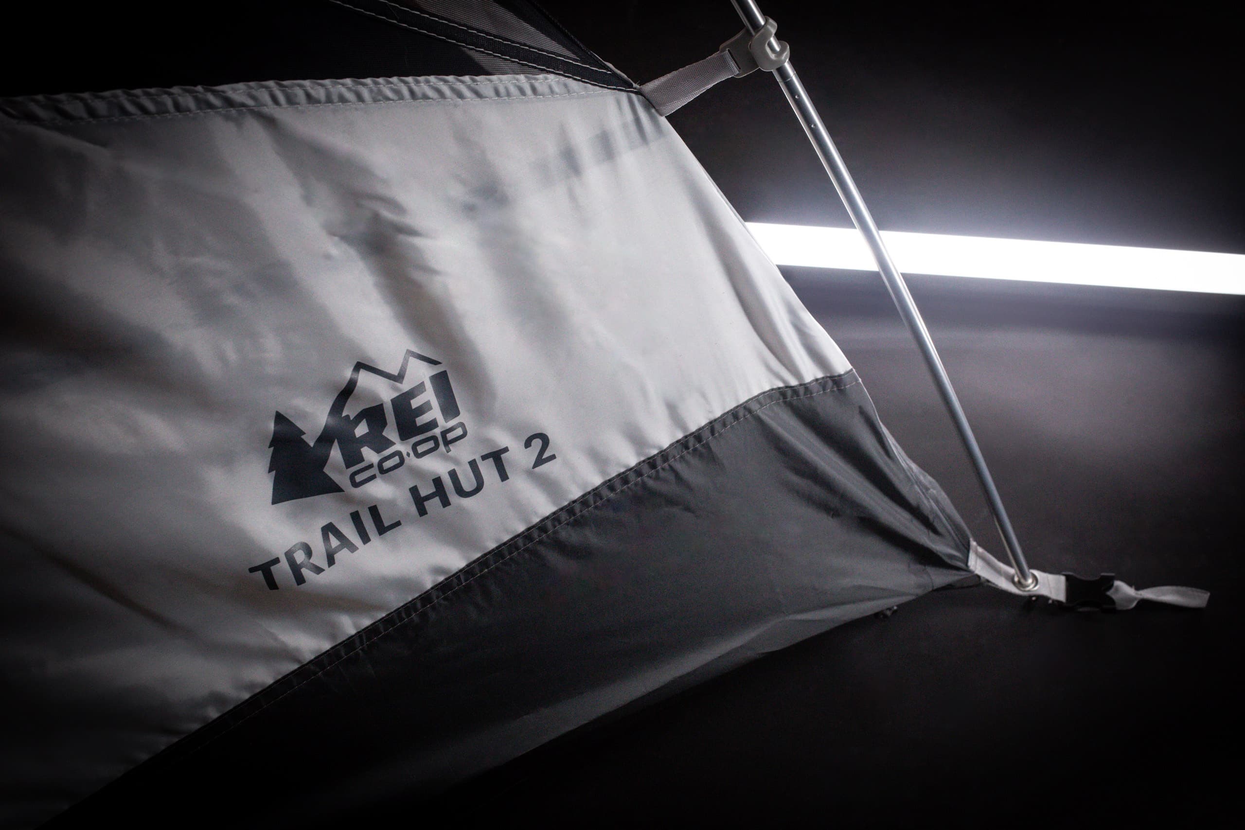 Close-up of a grey tent branded 'REI Co-op Trail Hut 2', with a pole and fabric detail visible against a dark background.