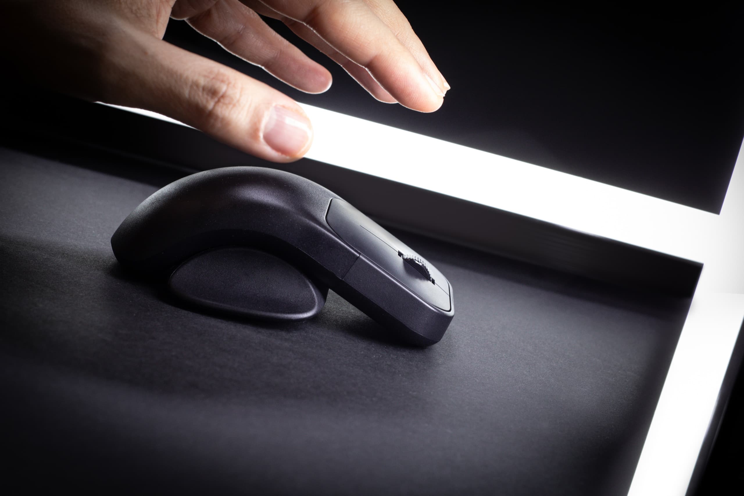 A hand poised to use a wireless ergonomic mouse on a dark surface, highlighted by a soft light.