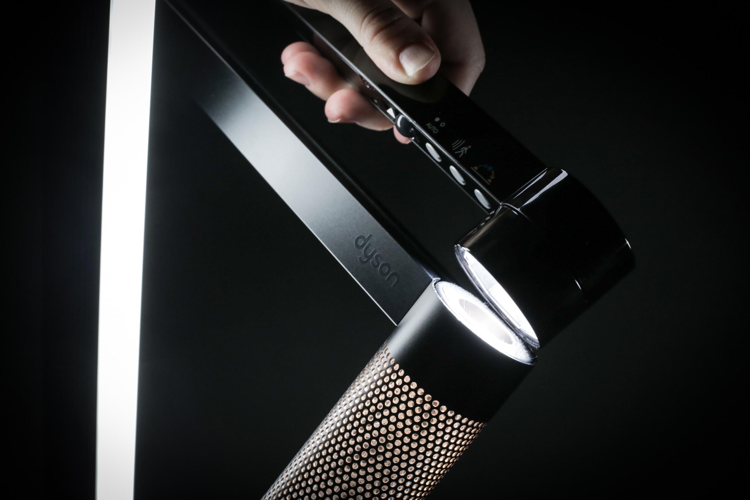 A hand adjusting a Dyson lamp with a lit LED light and control panel visible on a sleek black body.