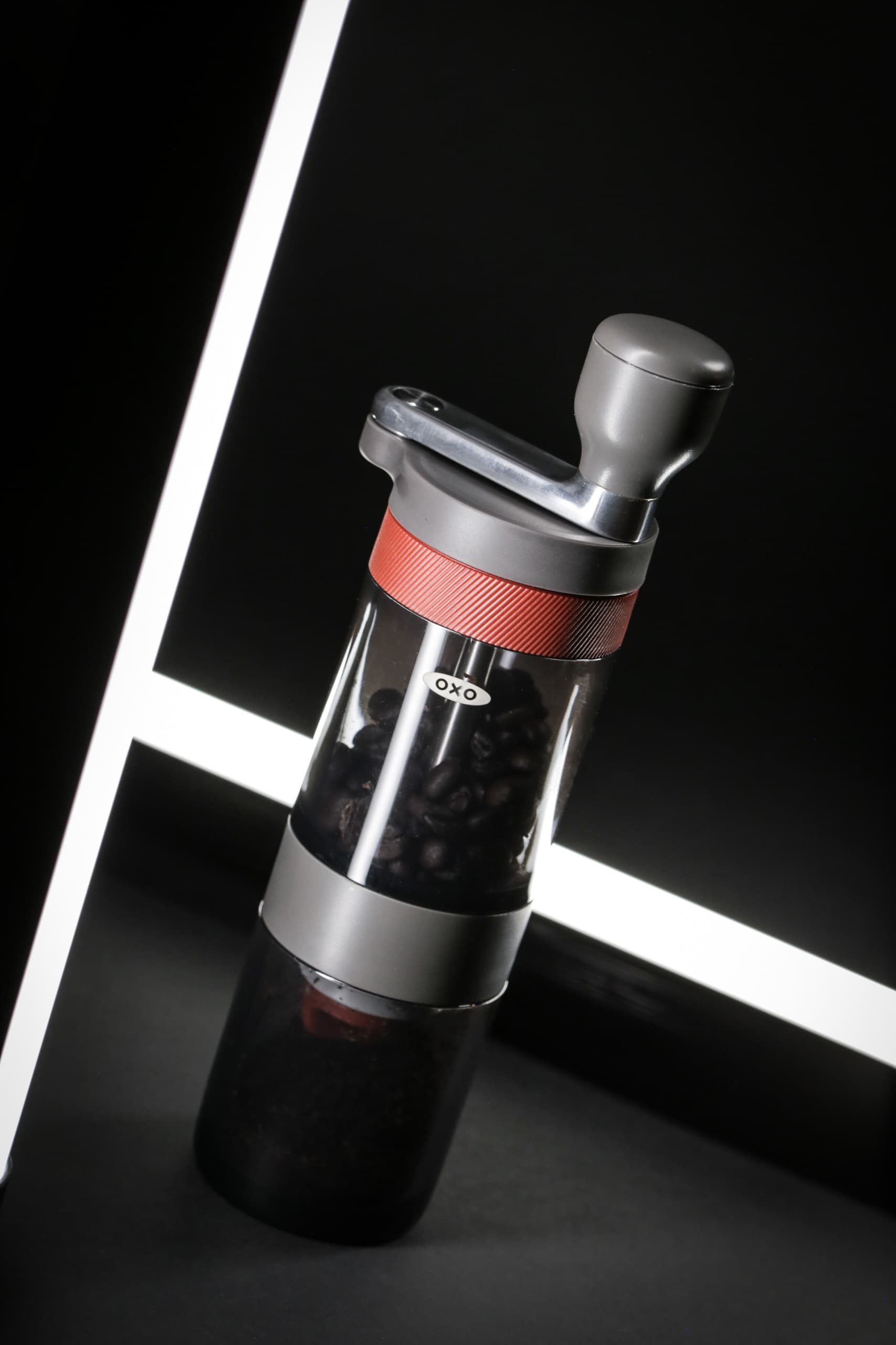 An OXO coffee grinder with a transparent container full of coffee beans, displayed on a dark surface beside a bright light strip.