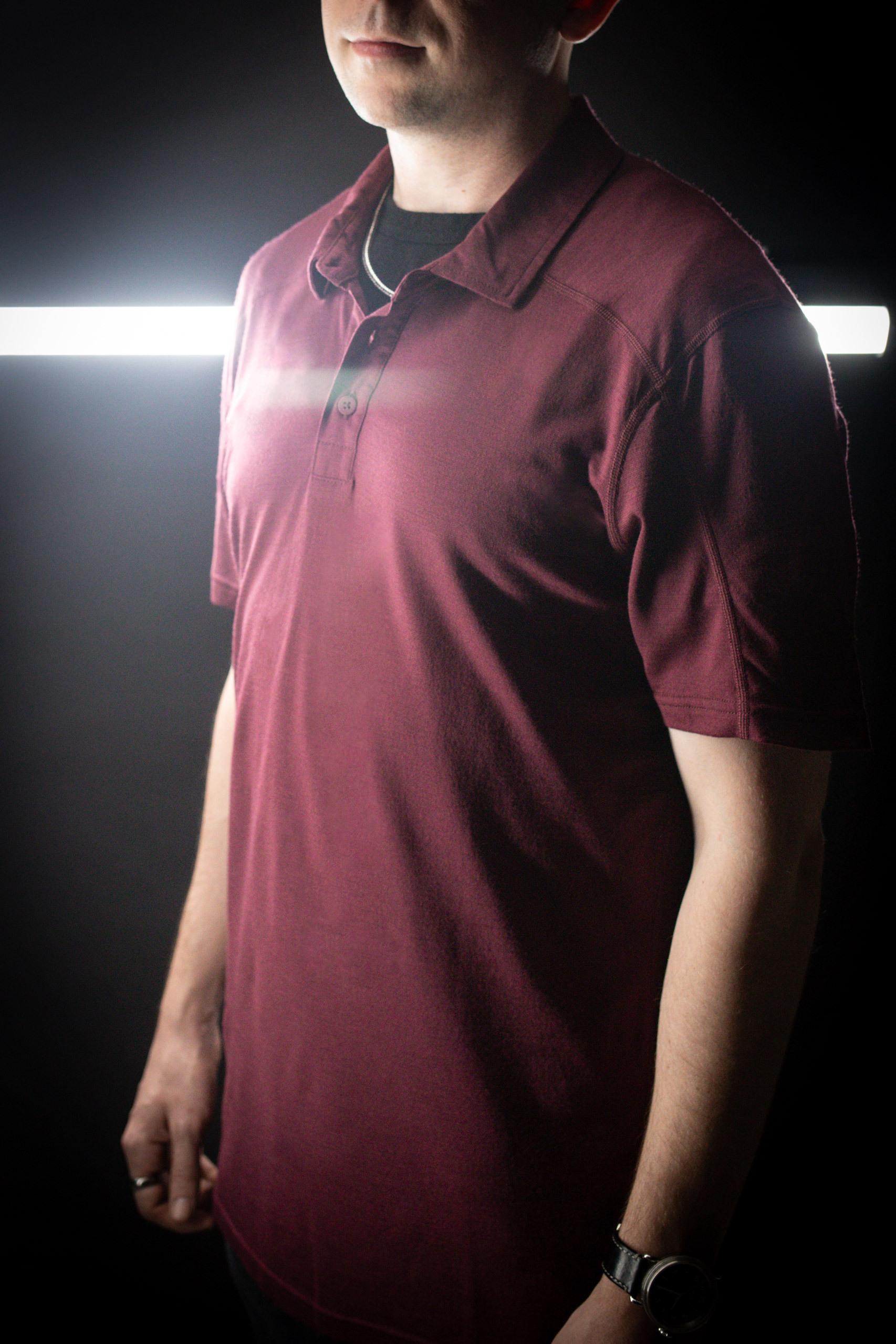 A man wearing a maroon polo shirt and a wristwatch, cropped at the shoulders, standing against a dark background with a white light strip.