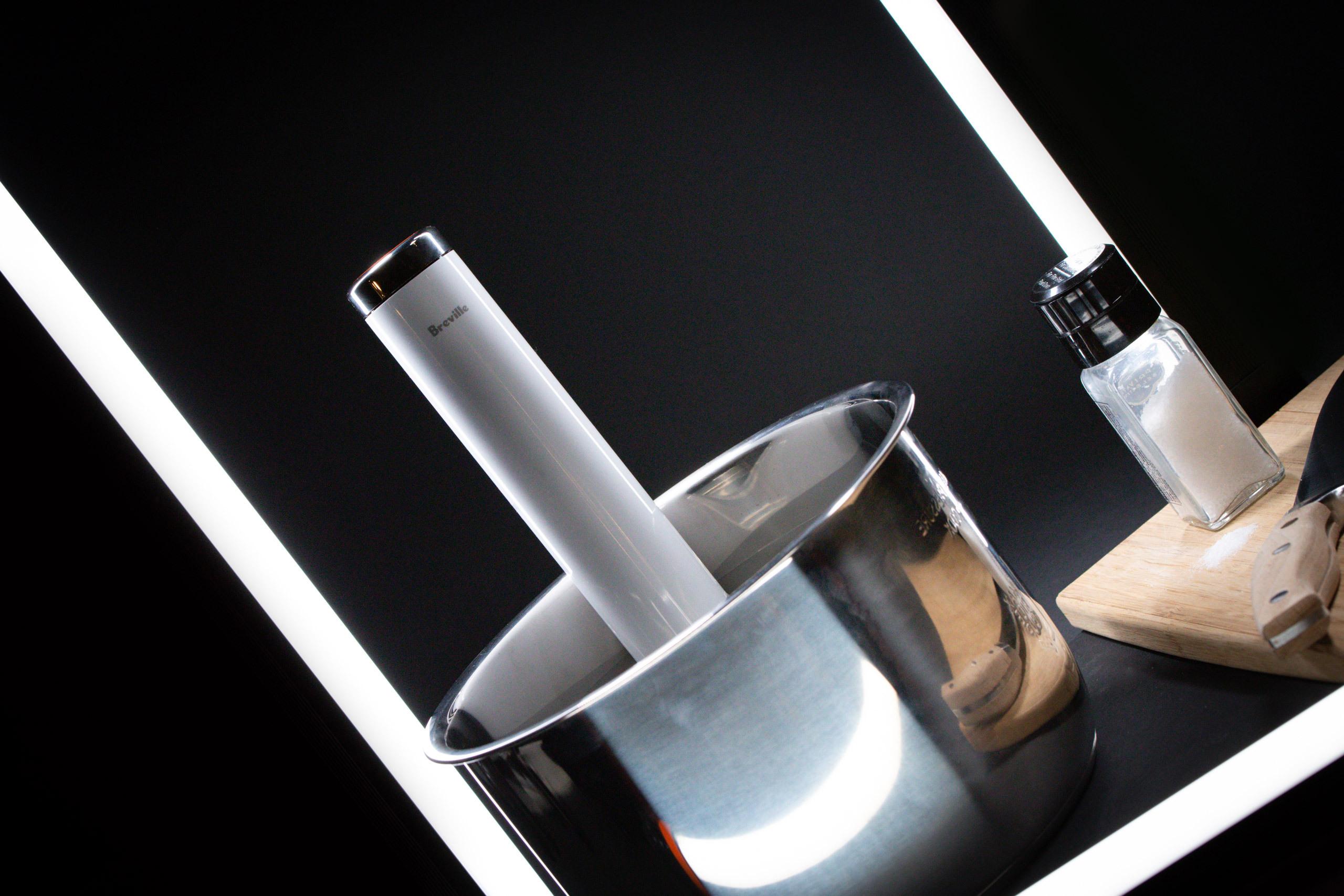 A stainless steel pot with an immersion blender inside, next to a salt shaker and wooden cutting board, illuminated by a white light strip.