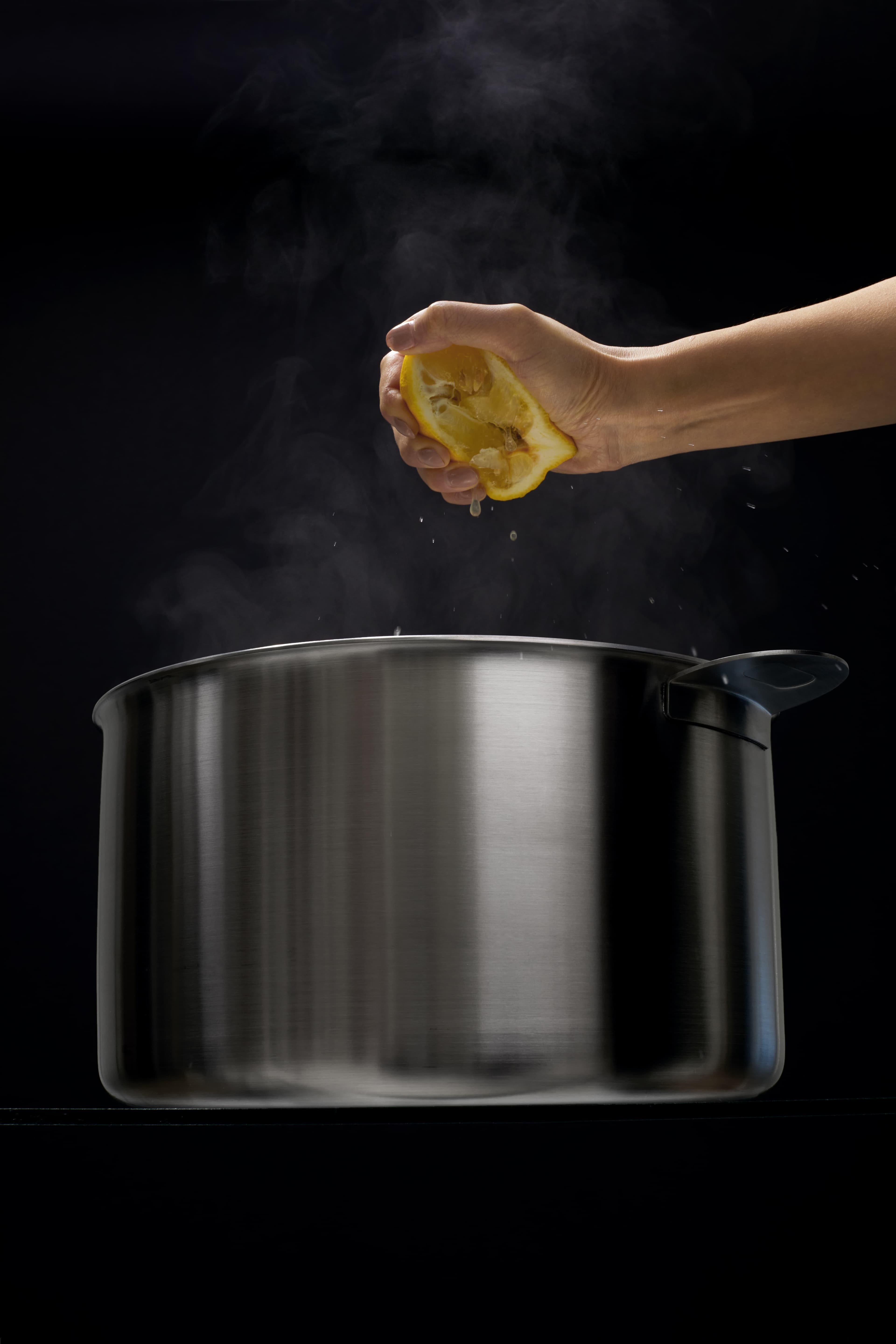 A hand squeezing a lemon half over a steaming stainless steel pot, with steam rising against a dark background.