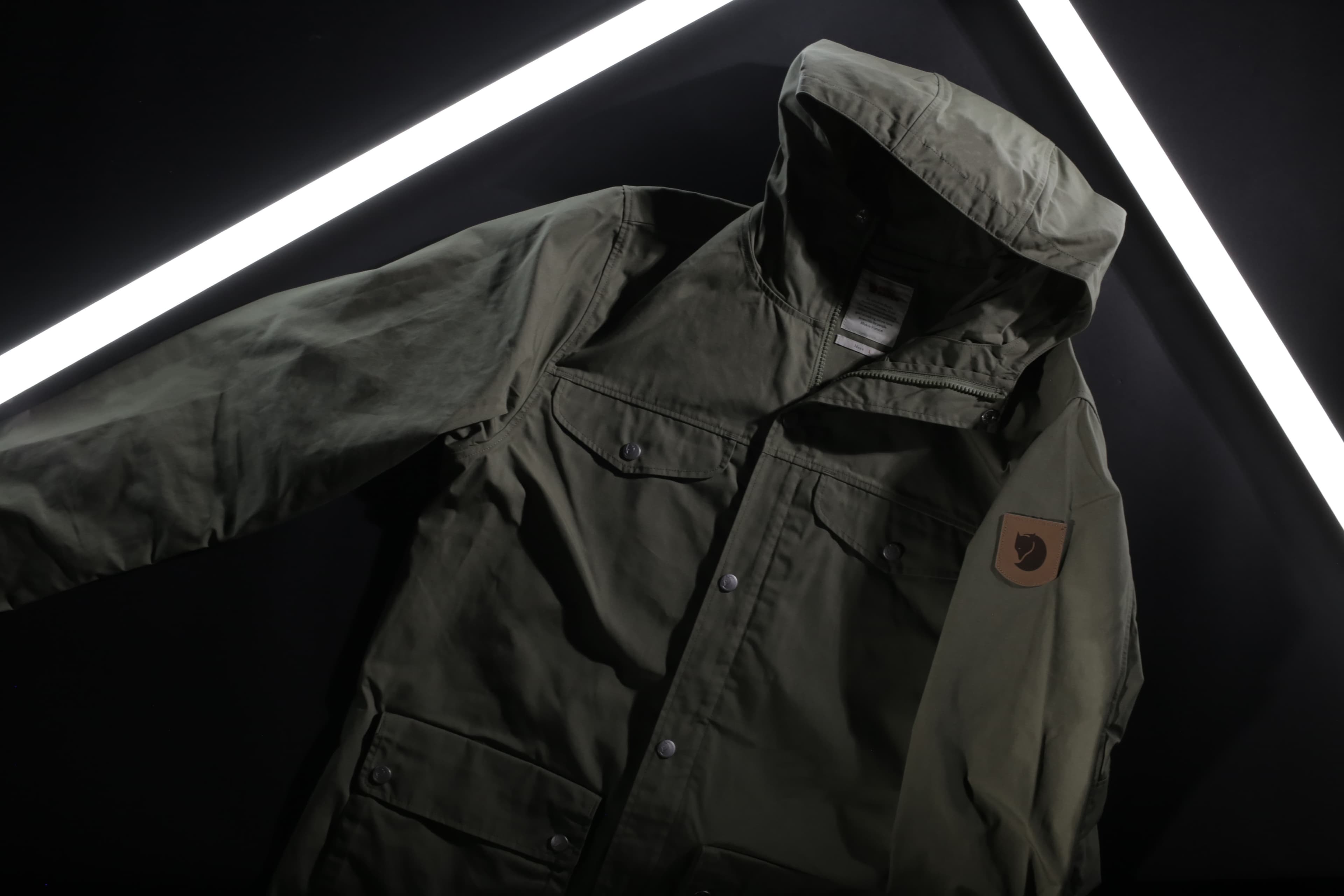 An olive green outdoor jacket with a hood and a logo patch on the sleeve, displayed in a dramatic lighting with two white light strips on a dark background.