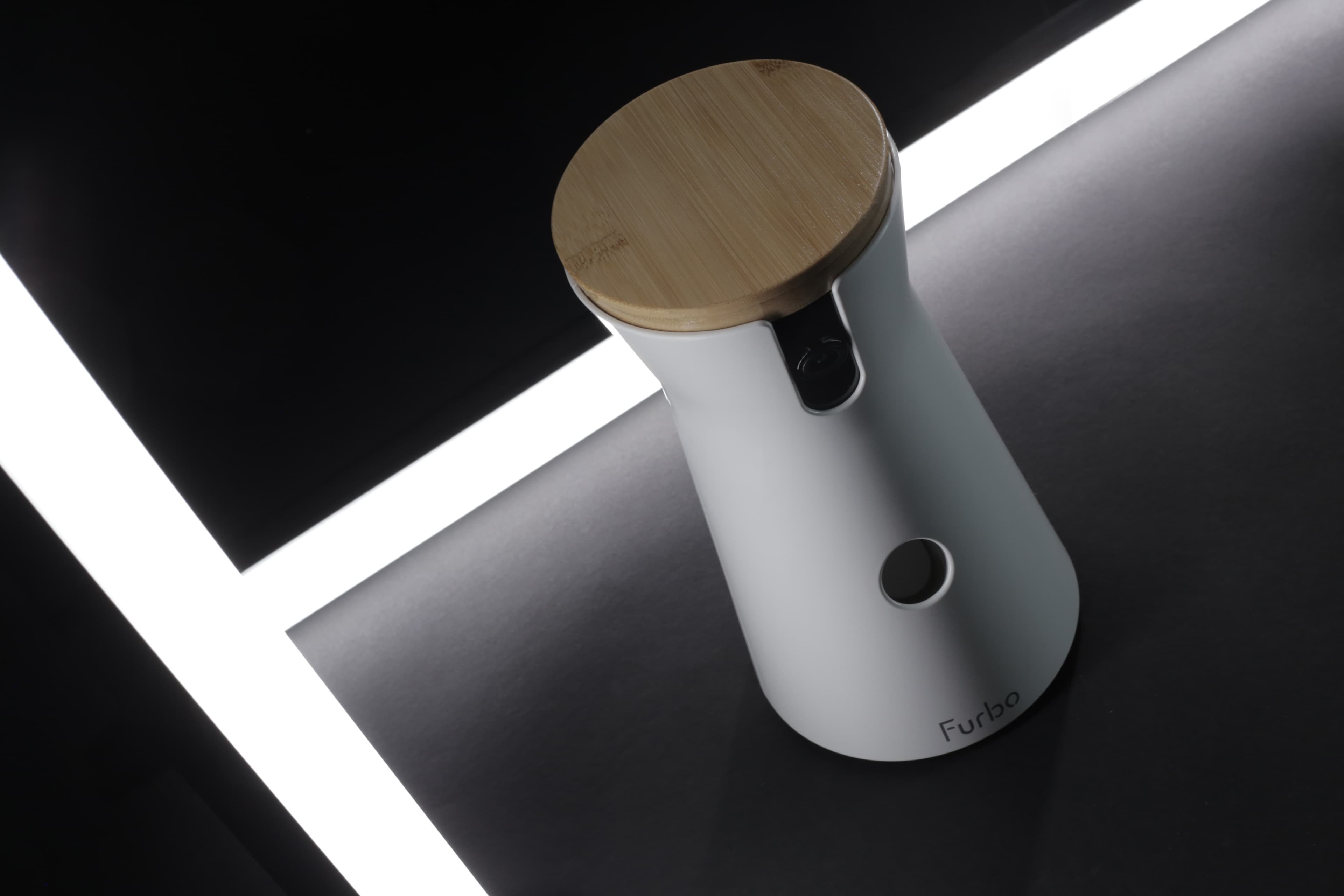 A modern Furbo dog camera with a wooden top and white body, displayed on a dark surface with surrounding light strips.