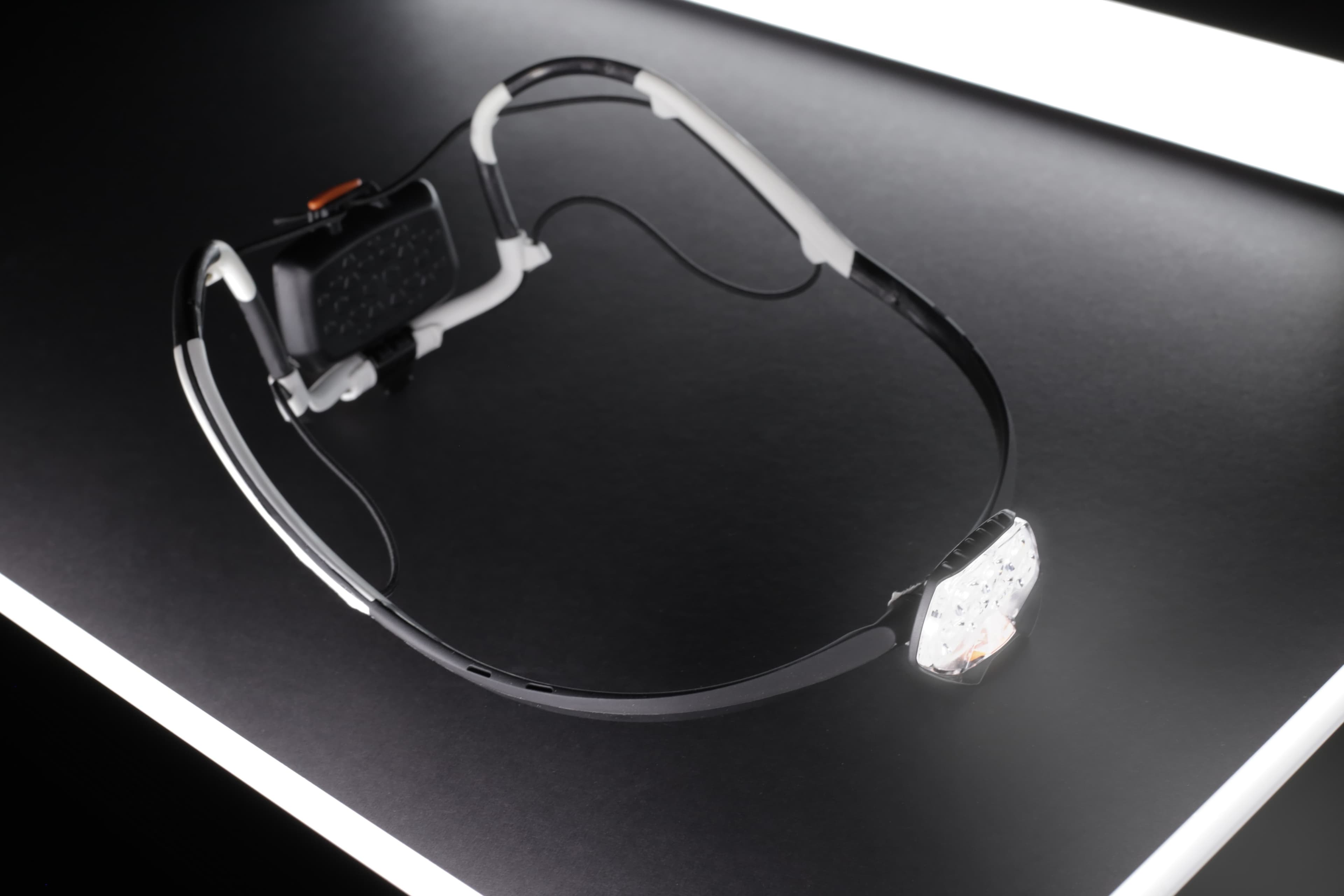 A high-tech head-mounted display lying on a dark surface, featuring transparent lenses and an attached electronics module, highlighted by parallel white light strips.