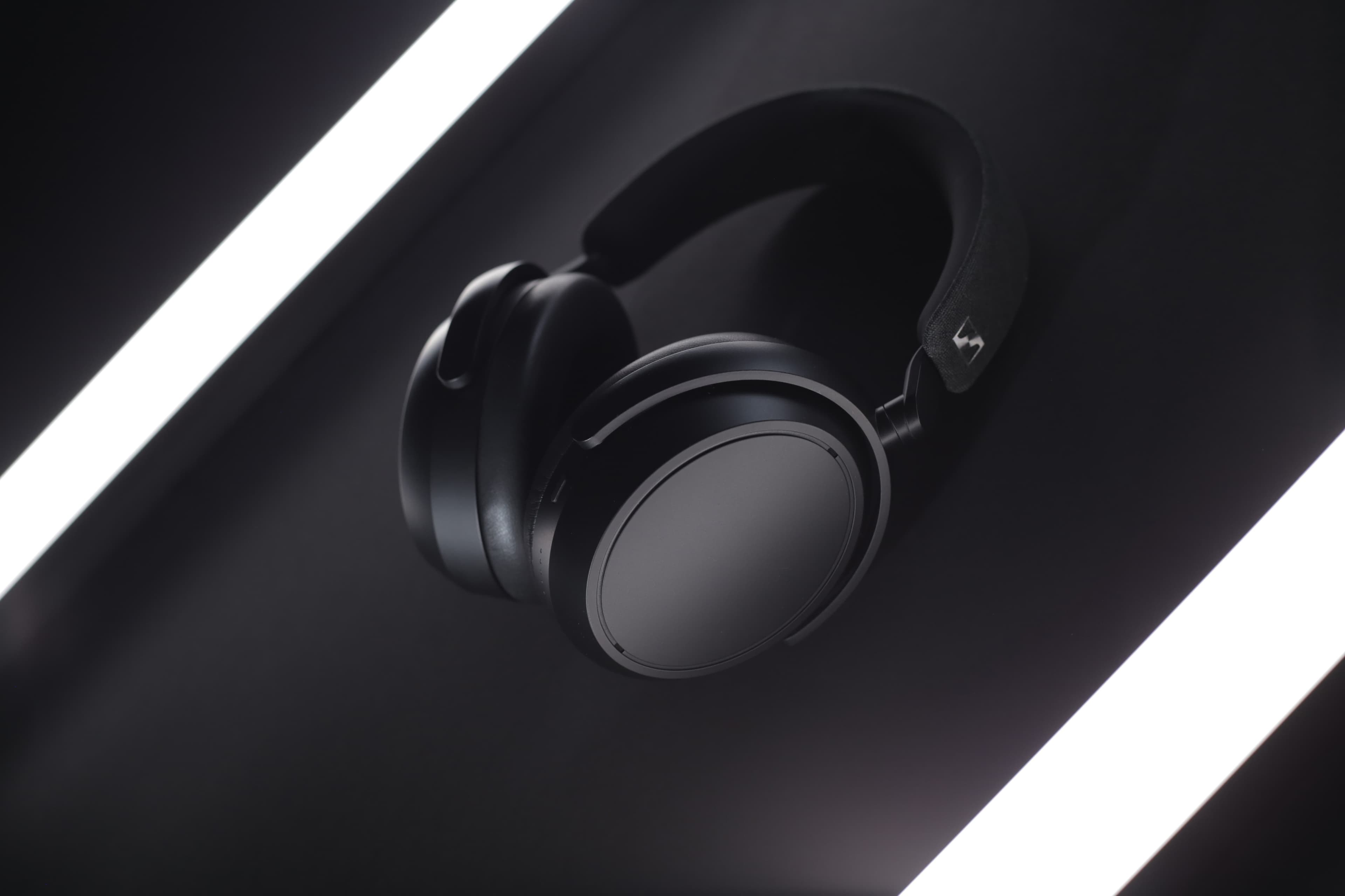 A pair of black over-ear headphones with a matte finish, set against a dark background with two bright light strips.