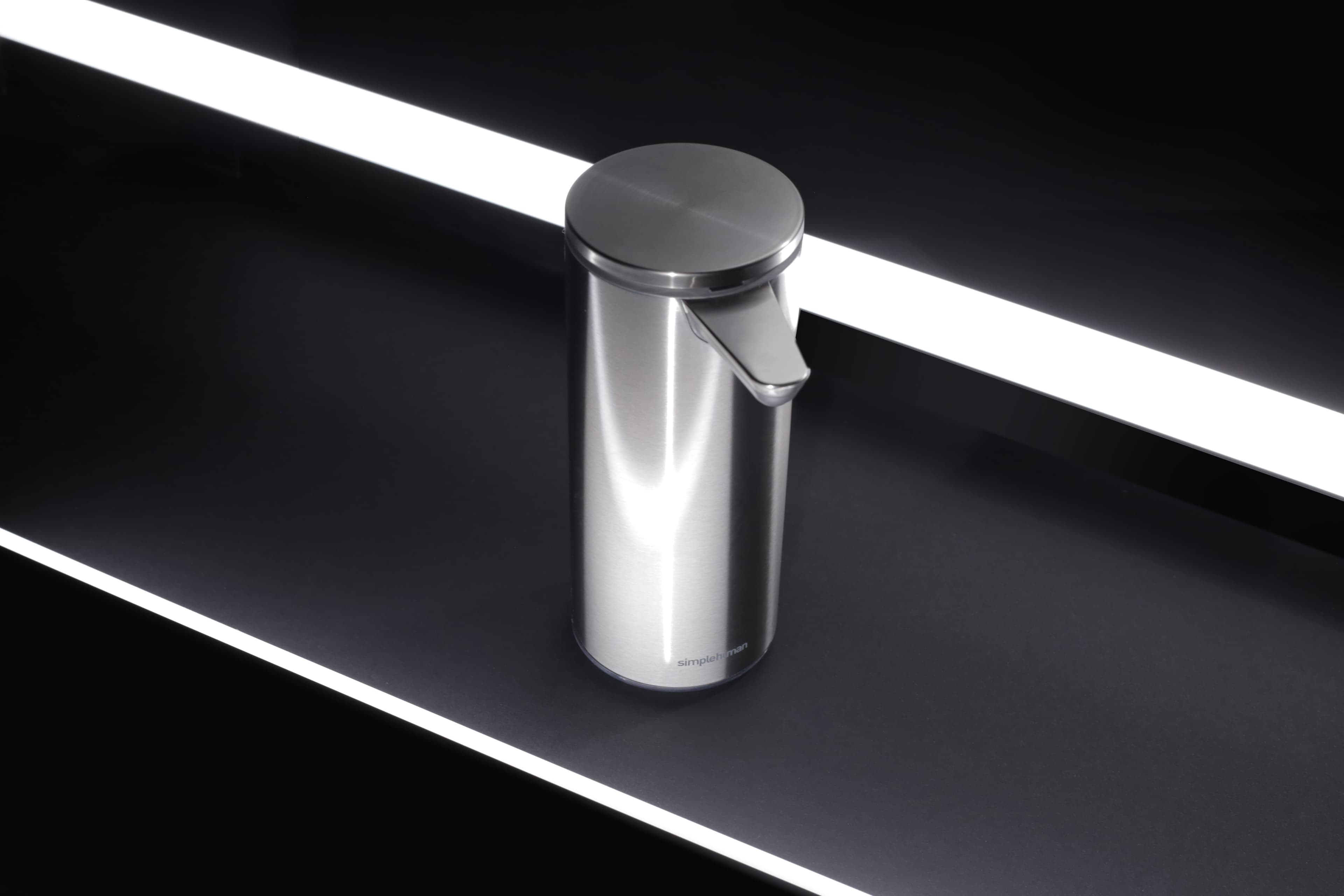 A stainless steel Simplehuman soap dispenser with a reflective surface, set against a dark background highlighted by two parallel white light strips.