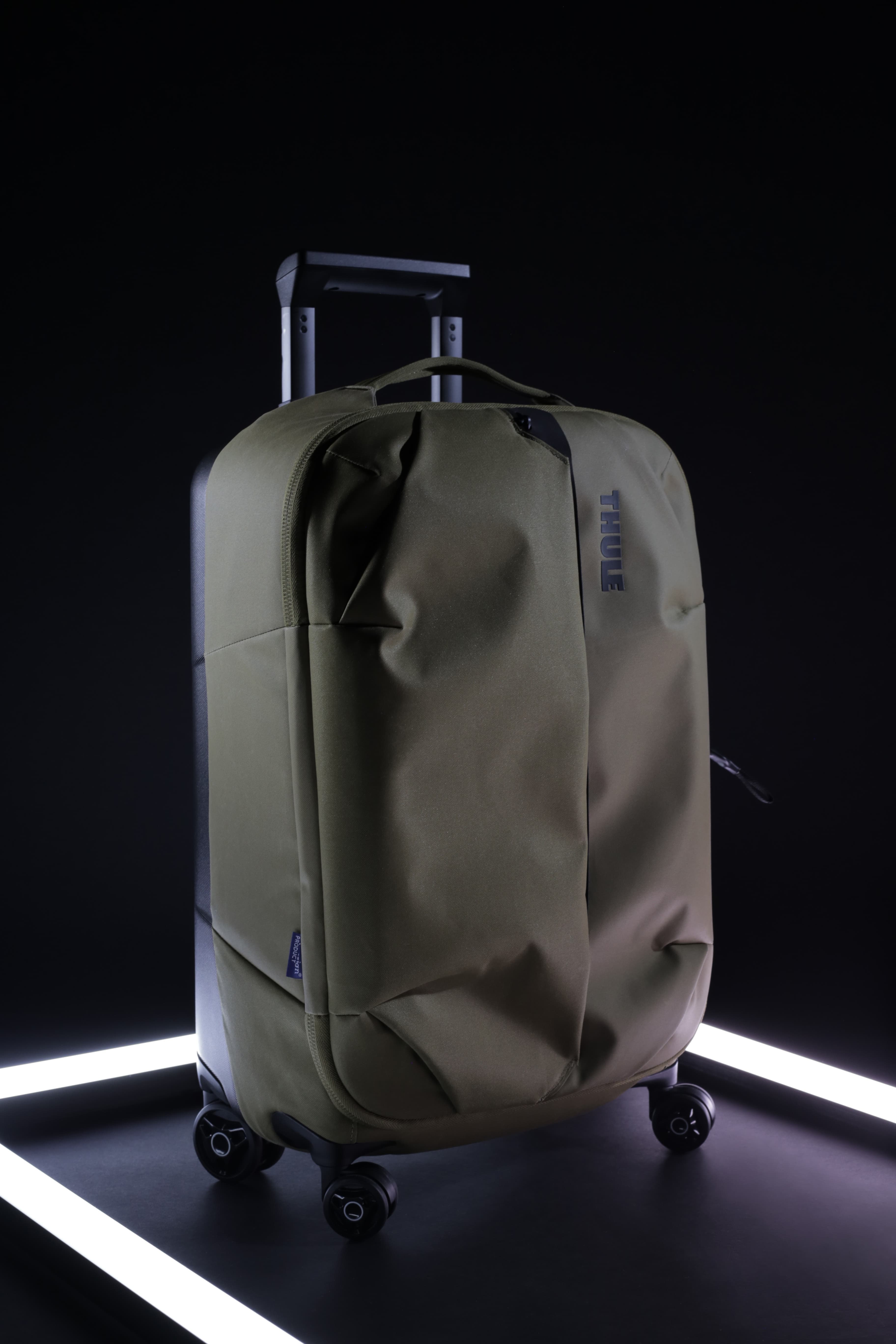 A Thule branded soft-sided olive green suitcase with an extendable handle and four wheels, set against a dark background with a white light circle.